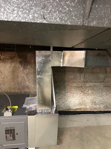 Metal ductwork connected to an electrical box and concrete wall, in a basement setting.