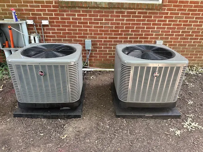 Two Rheem air conditioning units on black bases next to a brick wall.