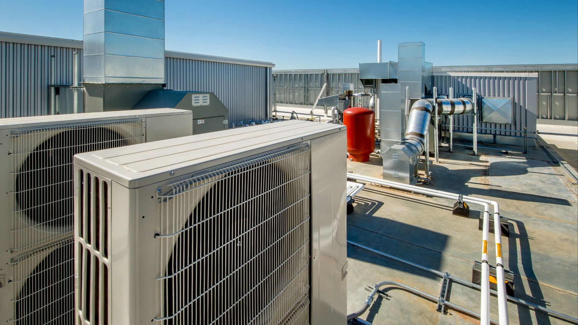 Rooftop with air conditioning units, ductwork, and a red tank under a blue sky.