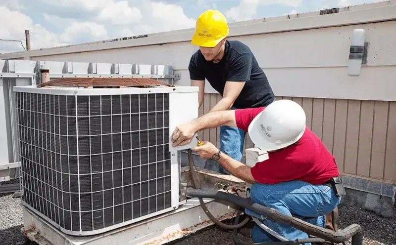 Two workers in hard hats kneel on a rooftop to repair an outdoor HVAC condenser unit.