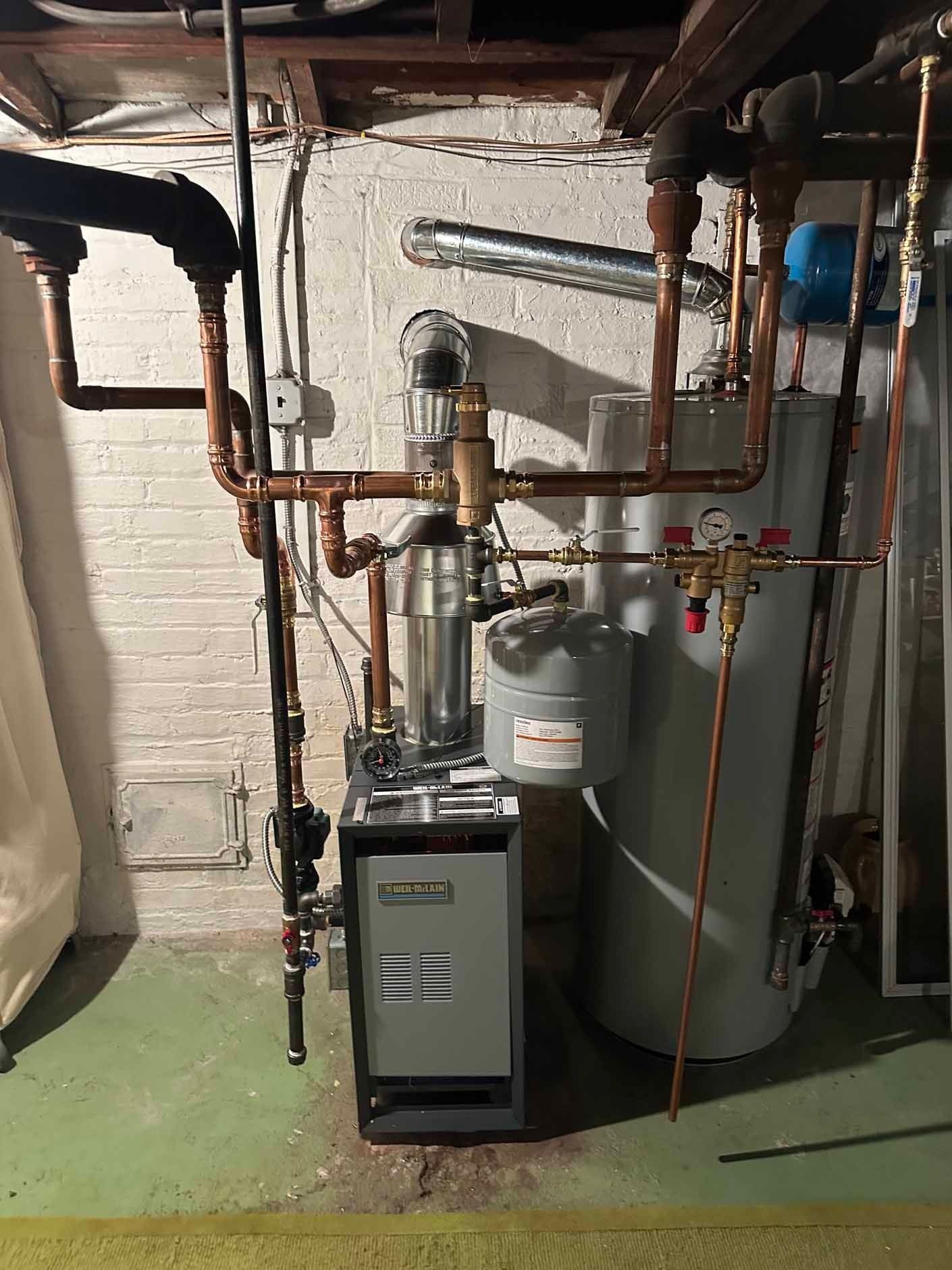 Furnace and water heater in a basement. Copper pipes and gray metal components against a white brick wall.