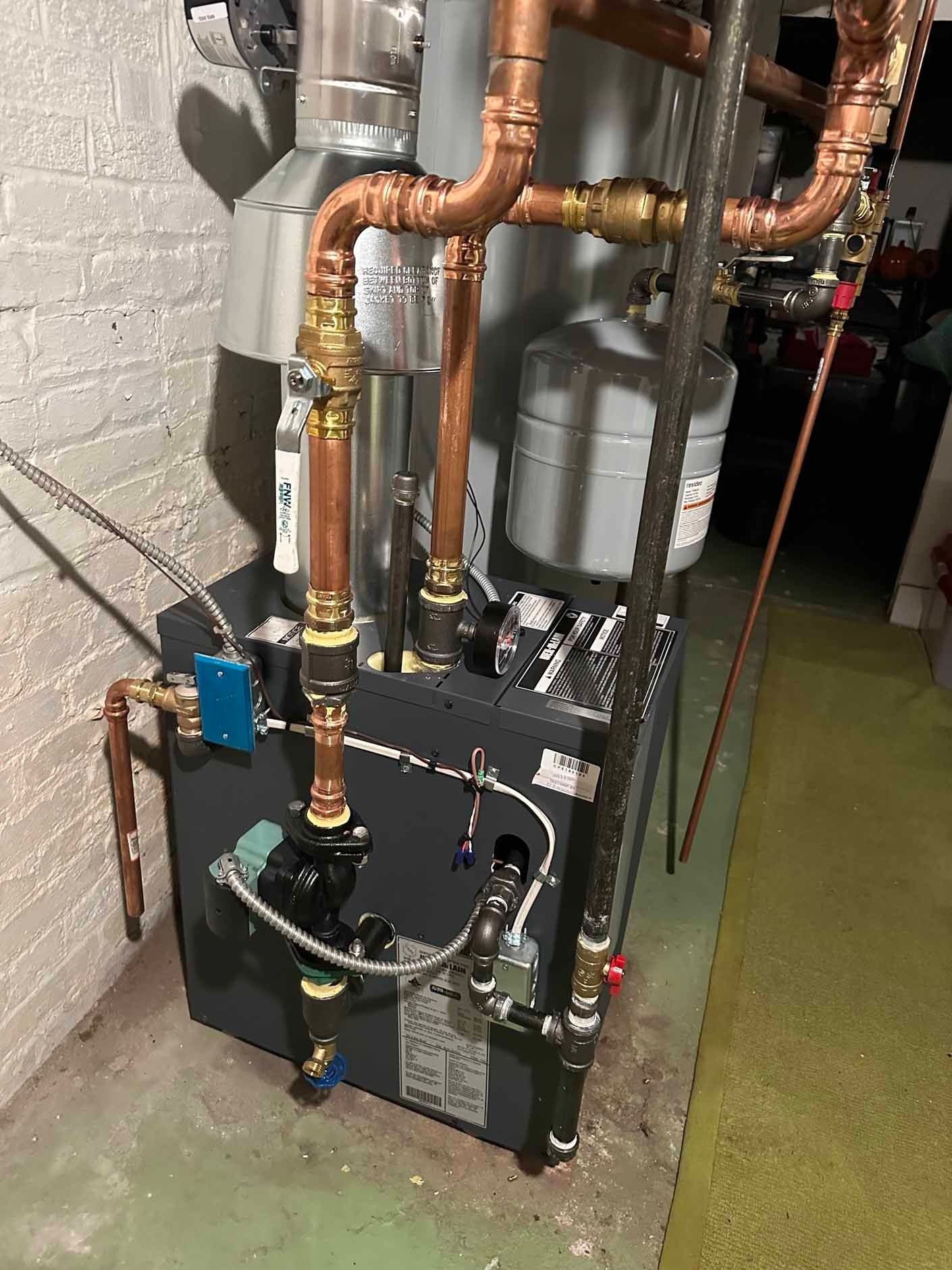 Heating system with copper pipes against a gray wall, including a silver expansion tank and black components.
