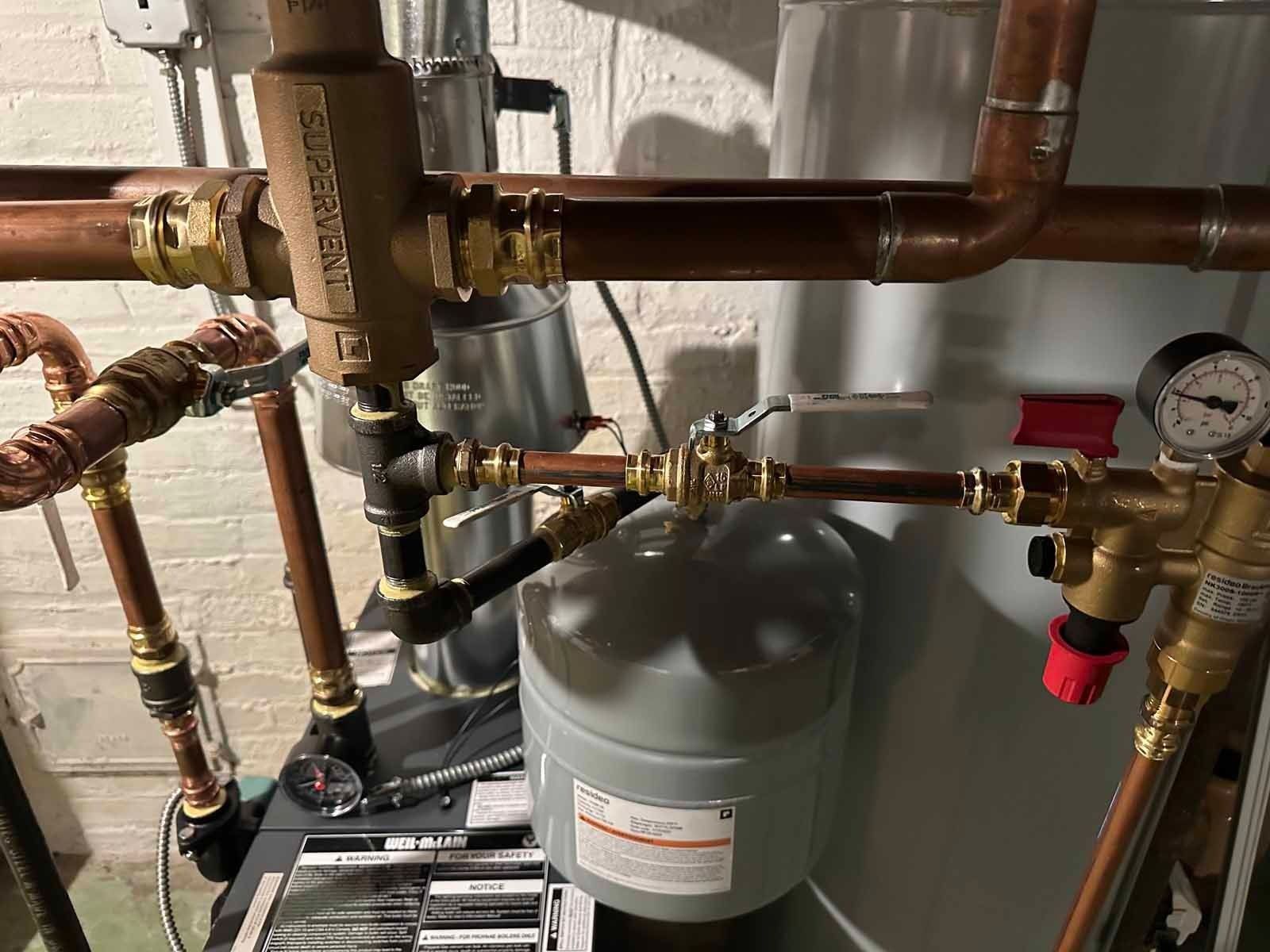 Copper pipes and valves connected to a water heater and expansion tank, set against a white wall.
