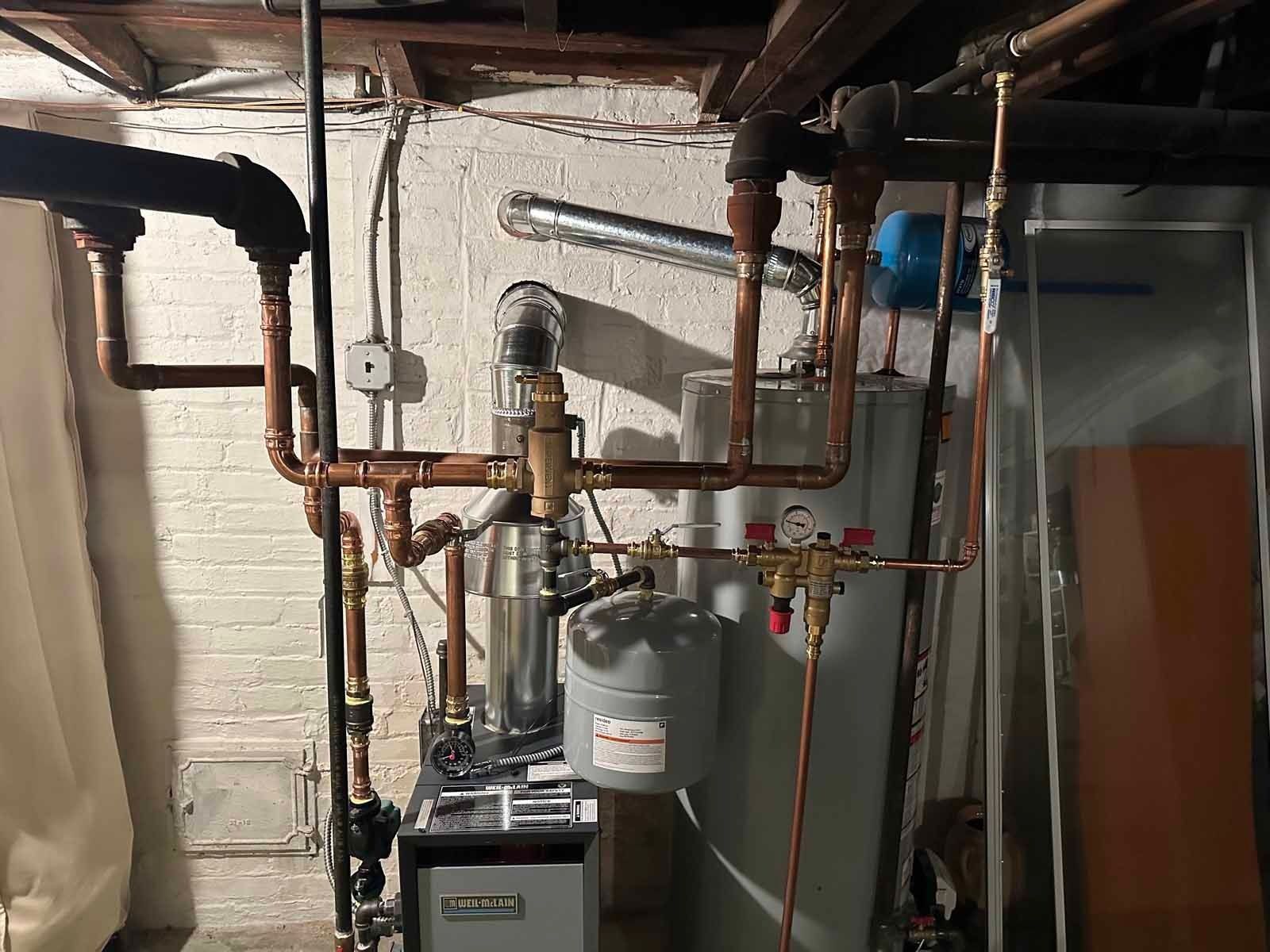 Copper pipes and a water heater in a basement, against a white wall, with a gray expansion tank.