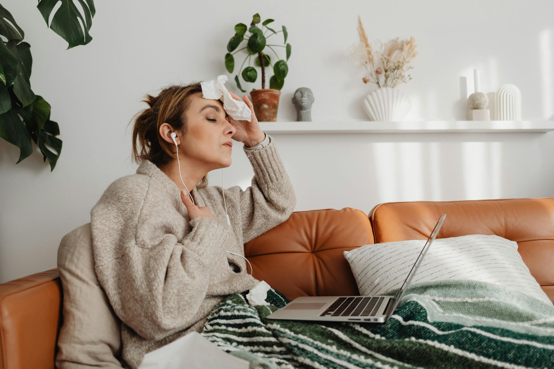 Woman on couch with cold, holding tissue to forehead, laptop nearby.