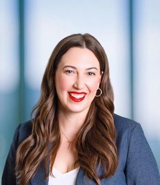 Smiling white woman in blue suit and white shirt with long brown hair, bright red lipstick and gold hoop earrings
