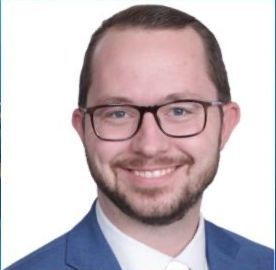 Smiling white man in blue suit and white shirt with short brown hair, beard, and brown glasses.