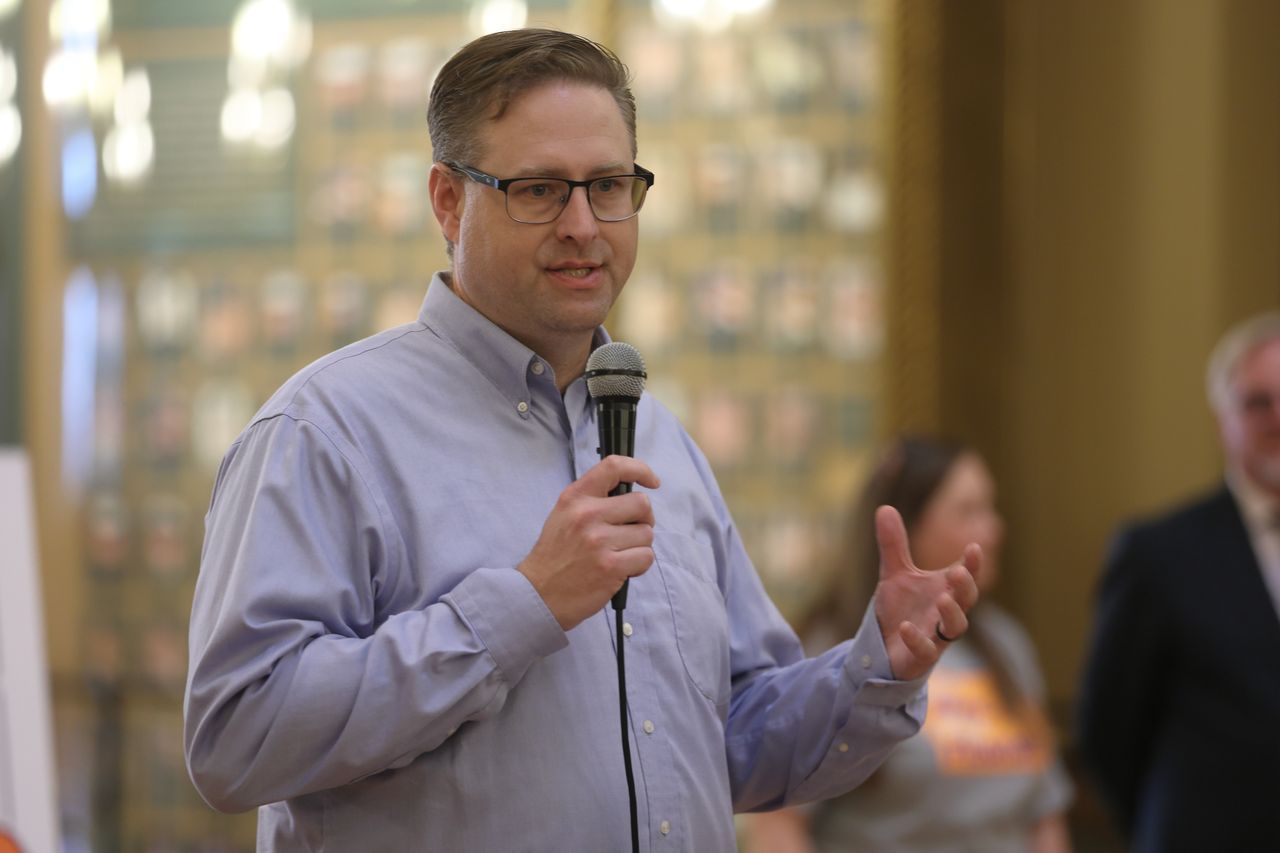 Photo of Kevin Harris speaking into a microphone to a crowd and gesturing with his hand. He has brown hair, is  wearing a lilac button-down shirt and glasses.