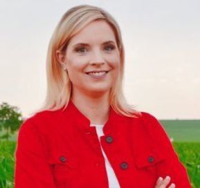 Photo of Ashley Hinson, blonde smiling white woman in a red jacket with Iowa landscape in backgound.