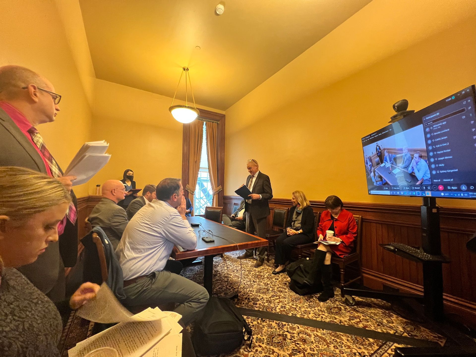 People sit around a table in a small room with others on zoom, listening to an advocate (Bill Stumpf) testify about guardianship law changes.