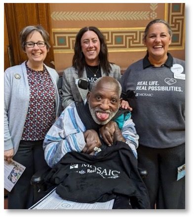 Picture of young black man in a wheelchair with big smile, holding a gray mosaic T-shirt with two staff behind him and Rep. Matson.