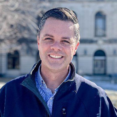 Photo of Zach Nunn, smiling dark haired white man with light blue shirt and dark blue jacket in front of a government building