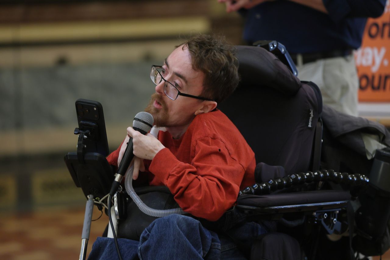 Man (Zach Mecham) in wheelchair with orange shirt, glasses, brown hair is holding a microphone and speaking to an audience at the Capitol. 