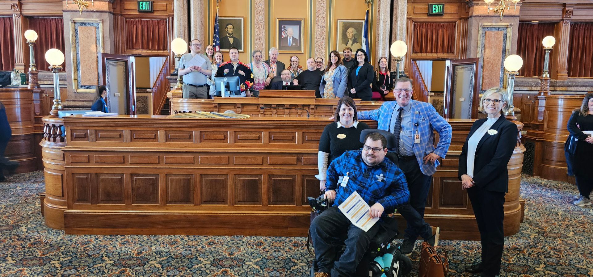 Photo of Speaker's seat in the Iowa House of Representatives with advocates and legislators gathered around. In the forefront is a man in a wheelchair and three others.