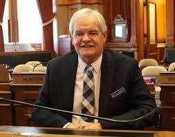 Rep. Graber smiling at his desk in Iowa House. He is a white man with white hair and mustache  wearing a blue suit with plaid tie.