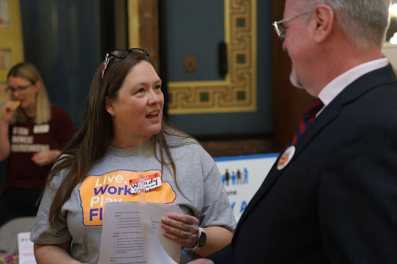 Wendy is wearing a gray shirt with Live.work. Play.Flourish written on it. She's holding a paper and speaking to an advocate who is in a black suit and red and blue stripped tie.
