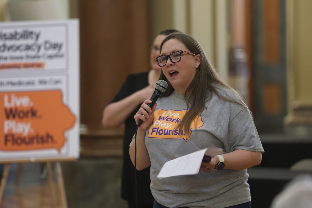 Wendy has long brown hair, wears glasses, and is wearing a gray shirt with an orange state of Iowa on it that says 