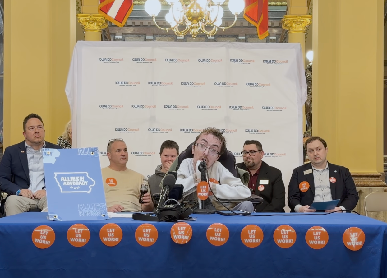 Zach, wearing a white shirt, sits in a wheelchair while speaking to the microphone at a panel held at the State Capitol. He is at a long table with a blue tablecloth and lots of orange stickers that say 