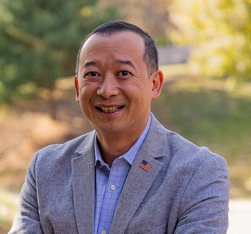 Photo of Lanon Baccam, a smiling Asian man in a light gray suit with outdoor scene in background