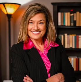 Photo of Sarah Corkery, a smiling white woman wearing a dark suit and bright pink blouse, arms crossed with bookshelf in background.