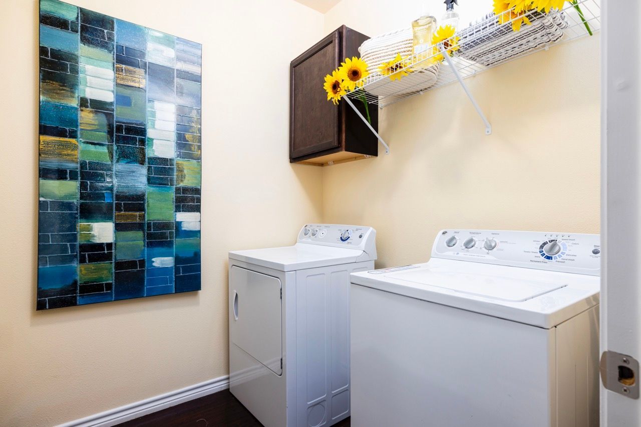 Compact apartment laundry room with a white washer and dryer, shelf above, and sunflowers.