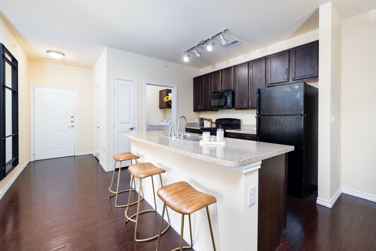 Open kitchen with granite island, dark wood cabinets, stainless appliances, and three bar stools.