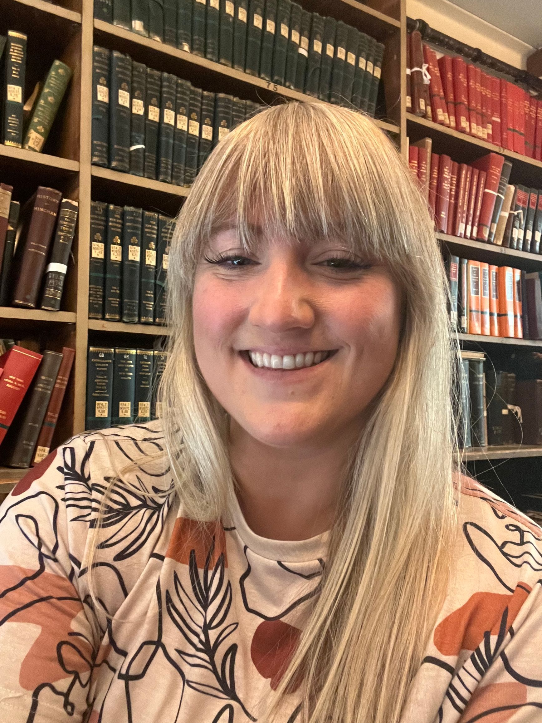 A woman is smiling in front of a bookshelf in a library.