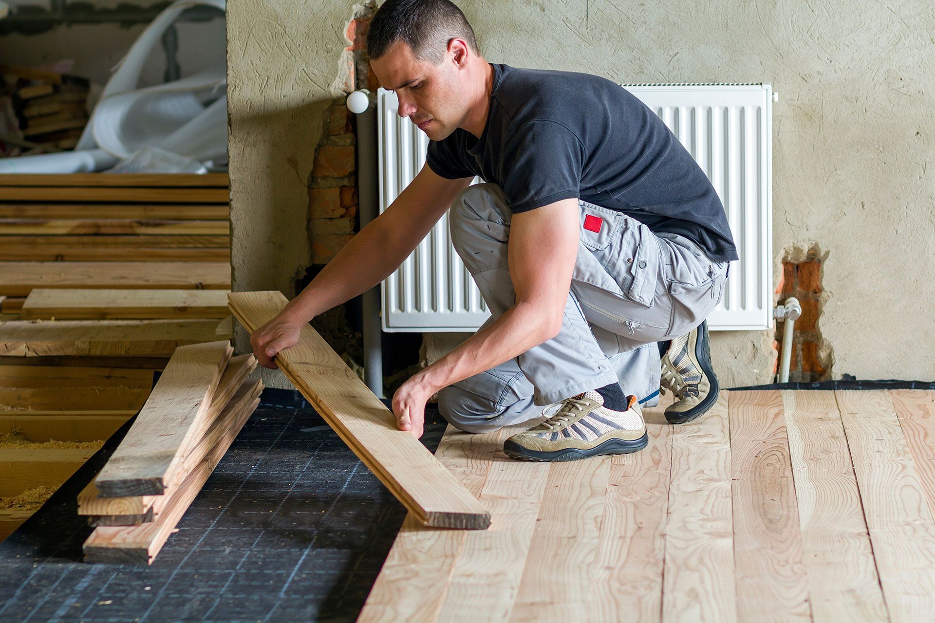 Young handsome professional carpenter installing natural wooden planks on wooden frame floor in empty unfinished room under reconstruction. Improvement, renovation and carpentry concept
