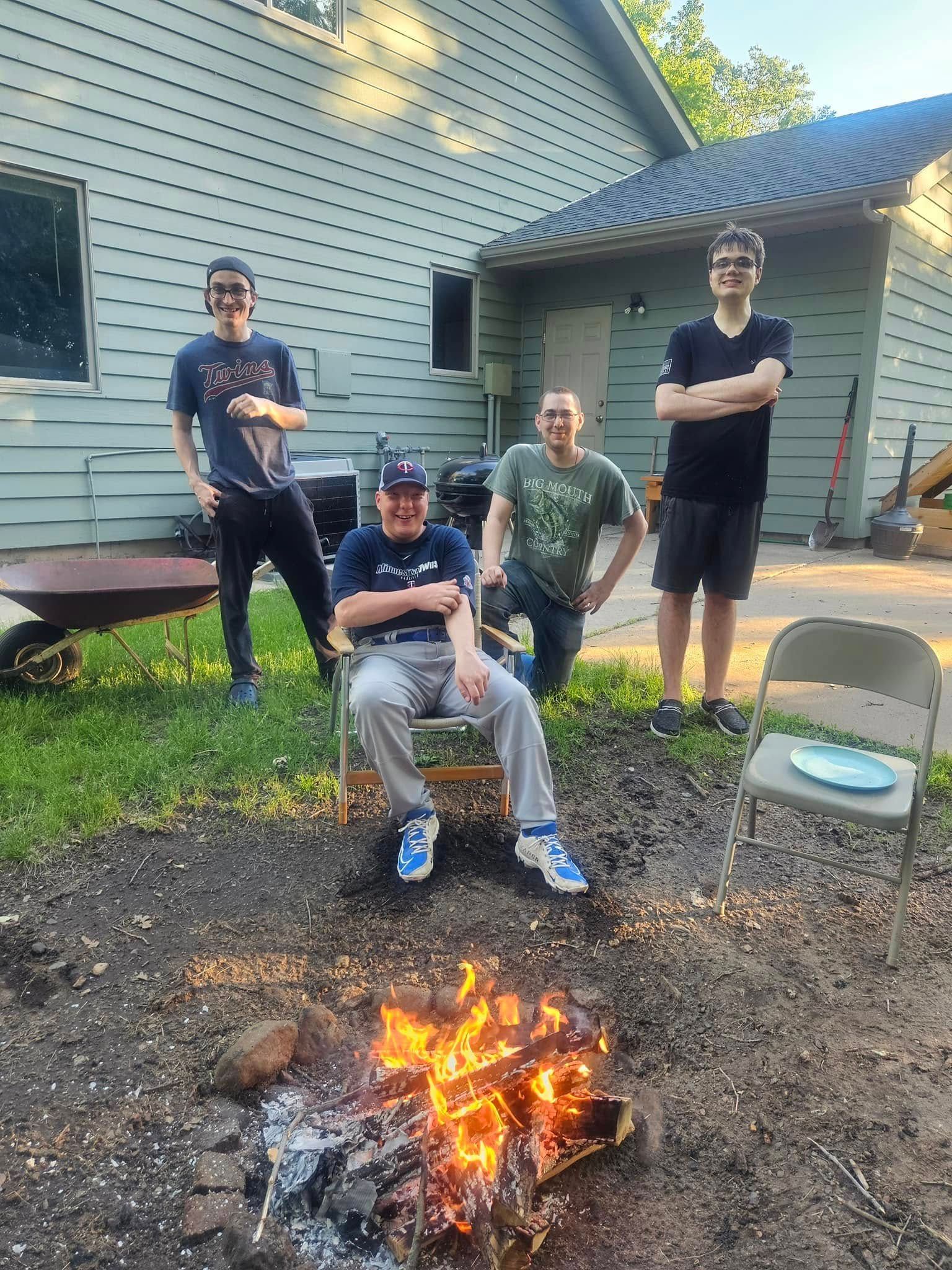 A group of men are sitting around a fire pit in front of a house.