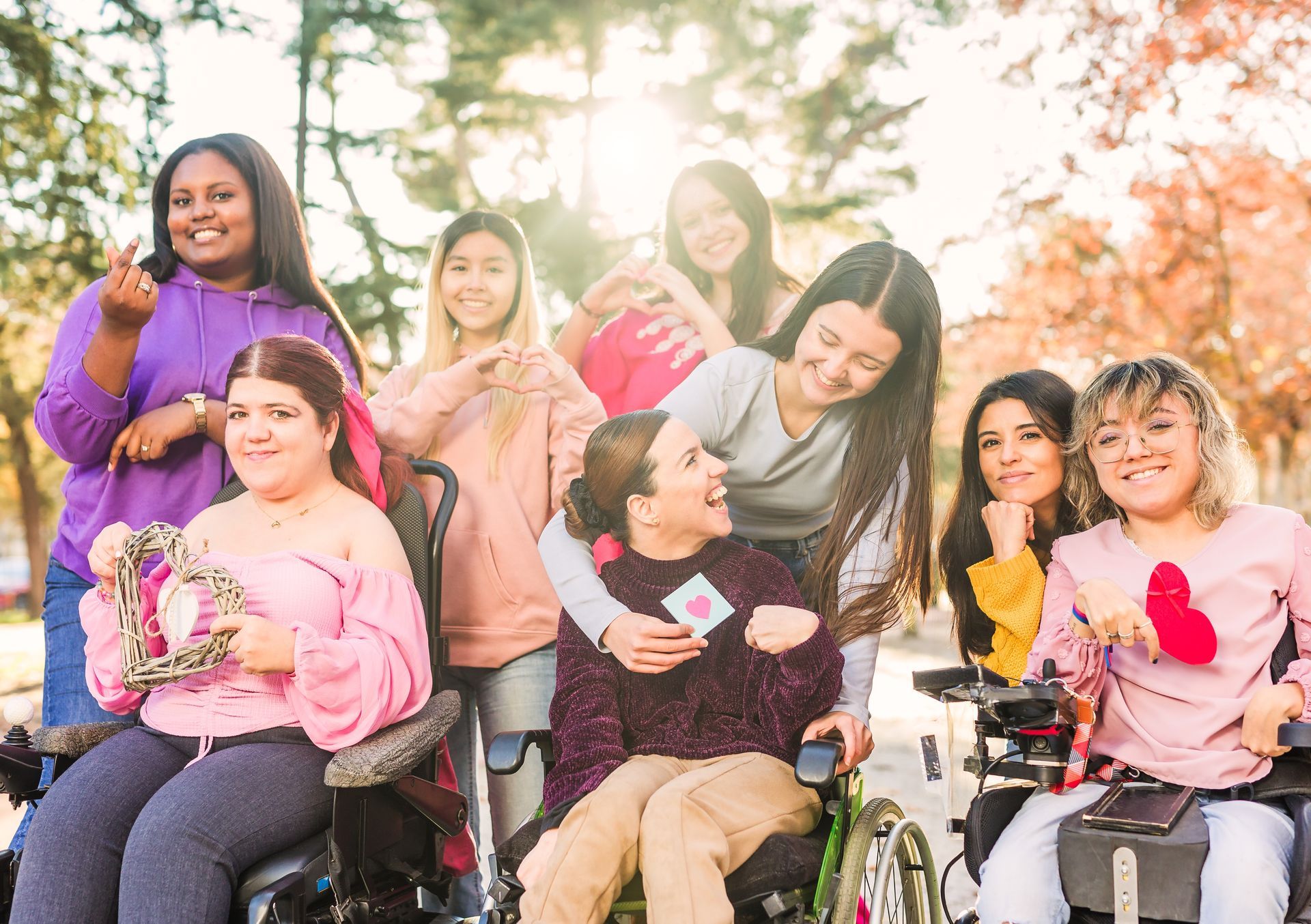 A group of diverse individuals, some in wheelchairs, smiling and holding heart-shaped objects in an outdoor park setting.