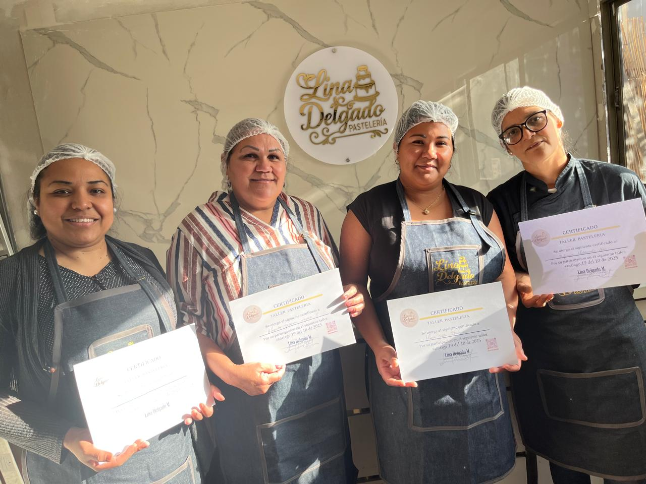 Cuatro personas con delantales sostienen certificados y sonríen en el interior, frente a un logotipo en la pared.