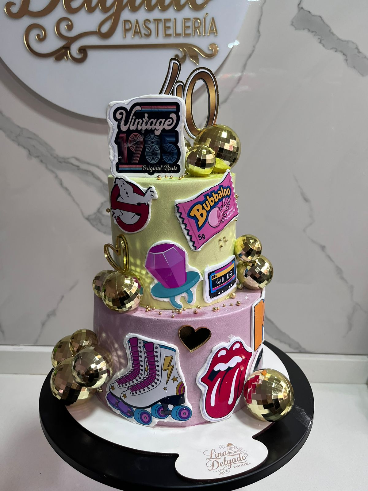 Tarta blanca de tres pisos con decoraciones de estilo cómic en rosa y dorado y esferas doradas sobre un soporte negro.