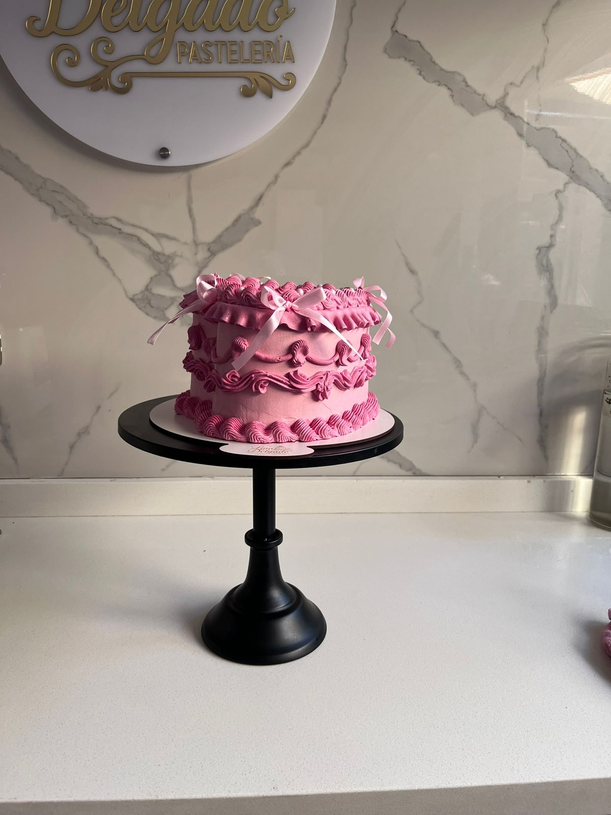 Pastel rosa glaseado sobre un pedestal negro contra una pared con estampado de mármol.