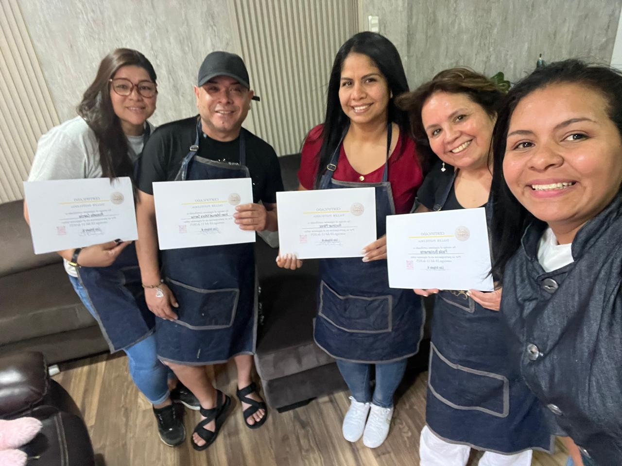 Cinco personas con delantales sonriendo y sosteniendo certificados en el interior de un edificio.