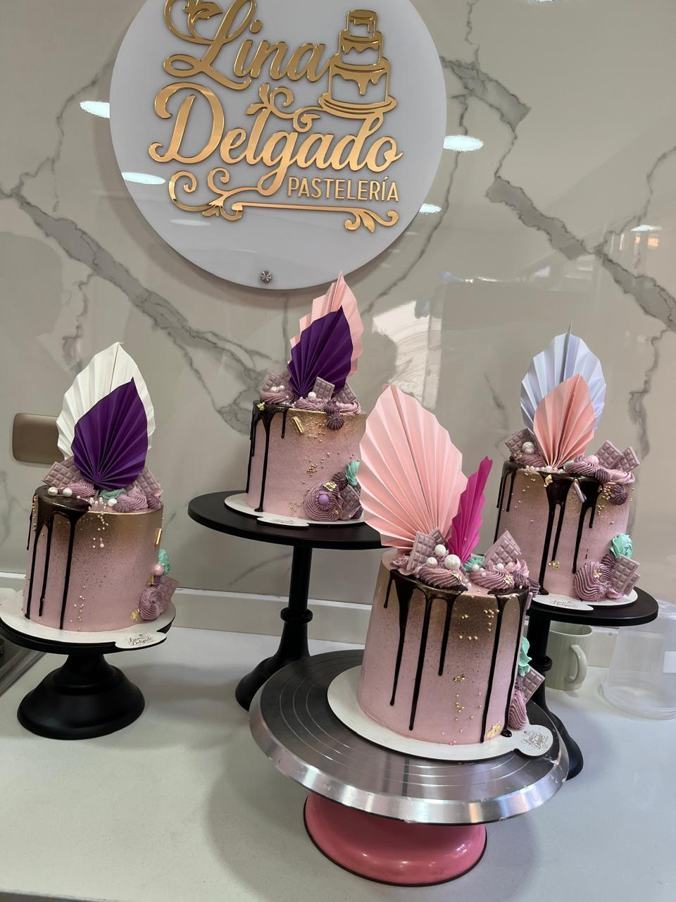 Cuatro pasteles decorados en rosa pastel con adornos de papel sobre soportes negros frente a un letrero personalizado.