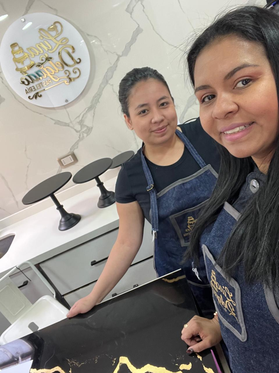 Dos personas con delantales posando en un espacio de trabajo de salón con mesas de manicura y herramientas.