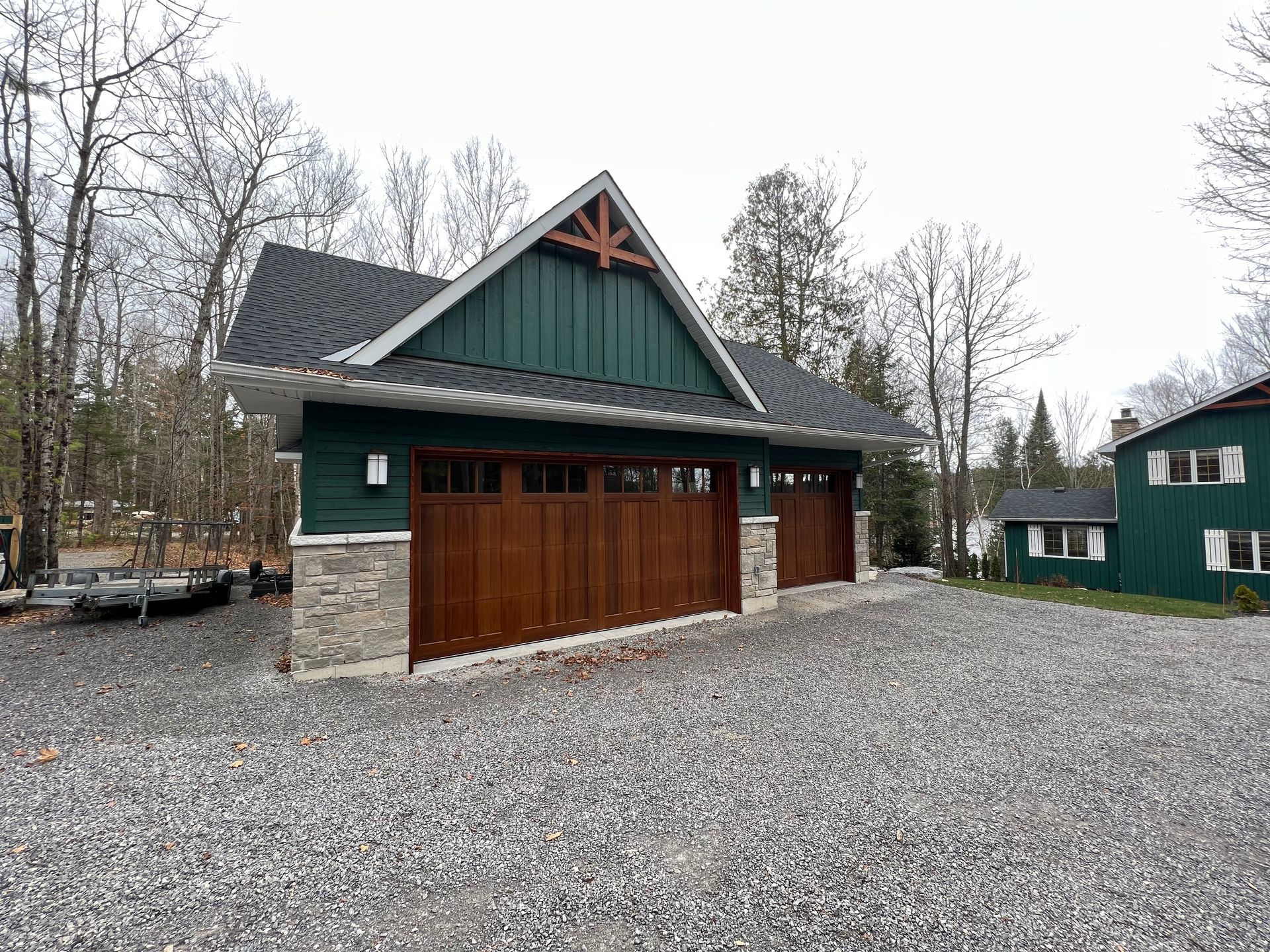 A garage with a green roof and wooden garage doors is sitting on top of a gravel driveway.