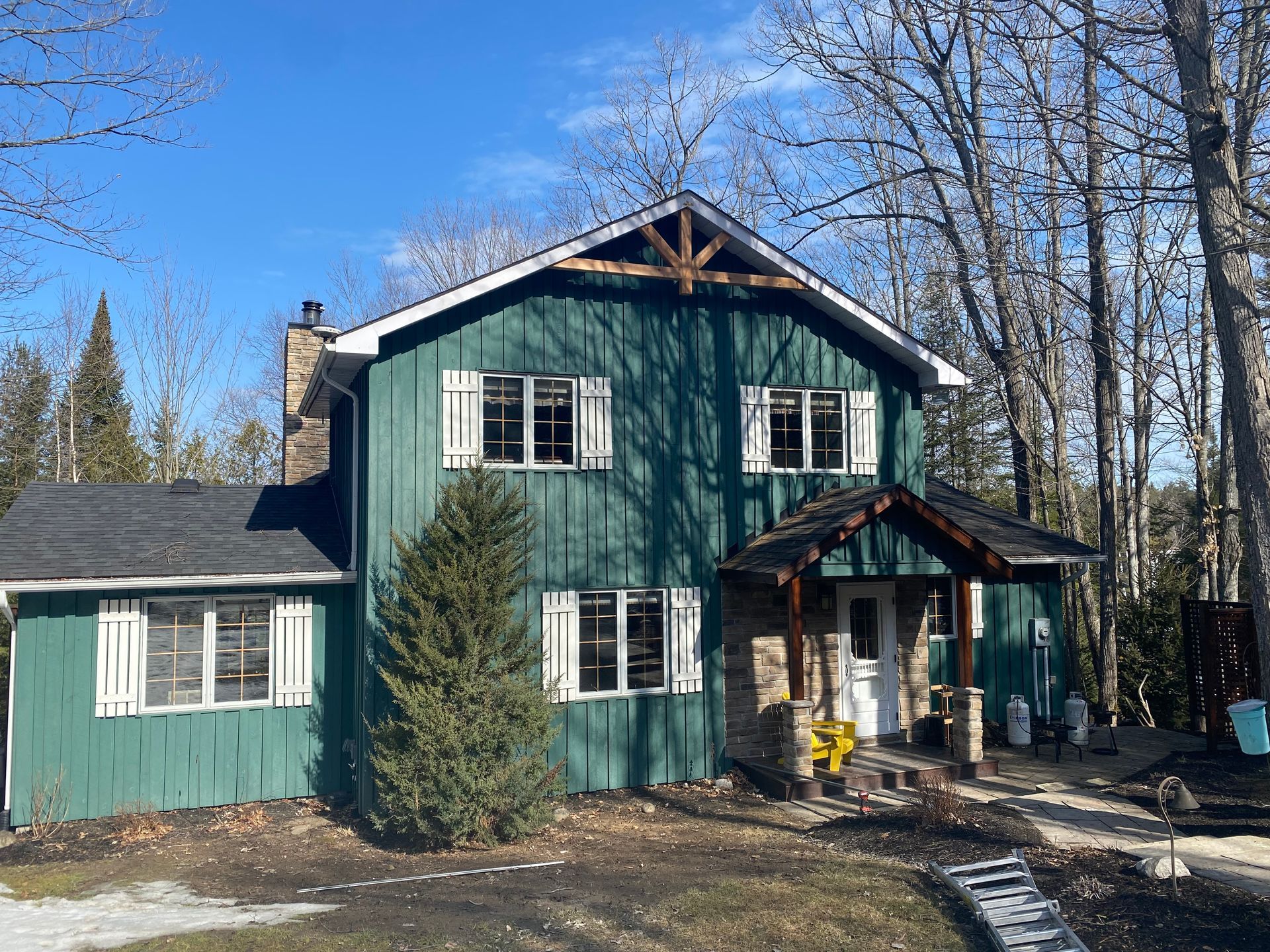 A green house with white shutters on the windows is surrounded by trees on a sunny day.