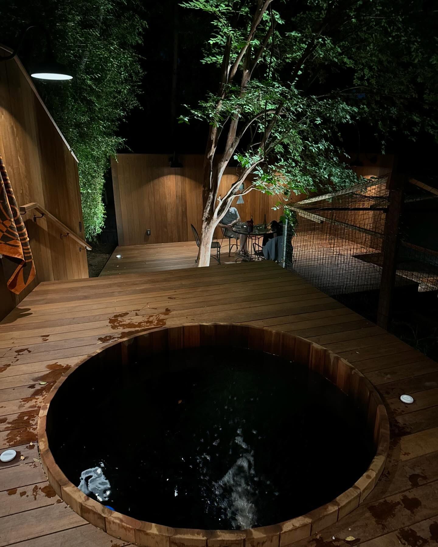 Wooden hot tub on a deck at night, with a tree and people in the background.