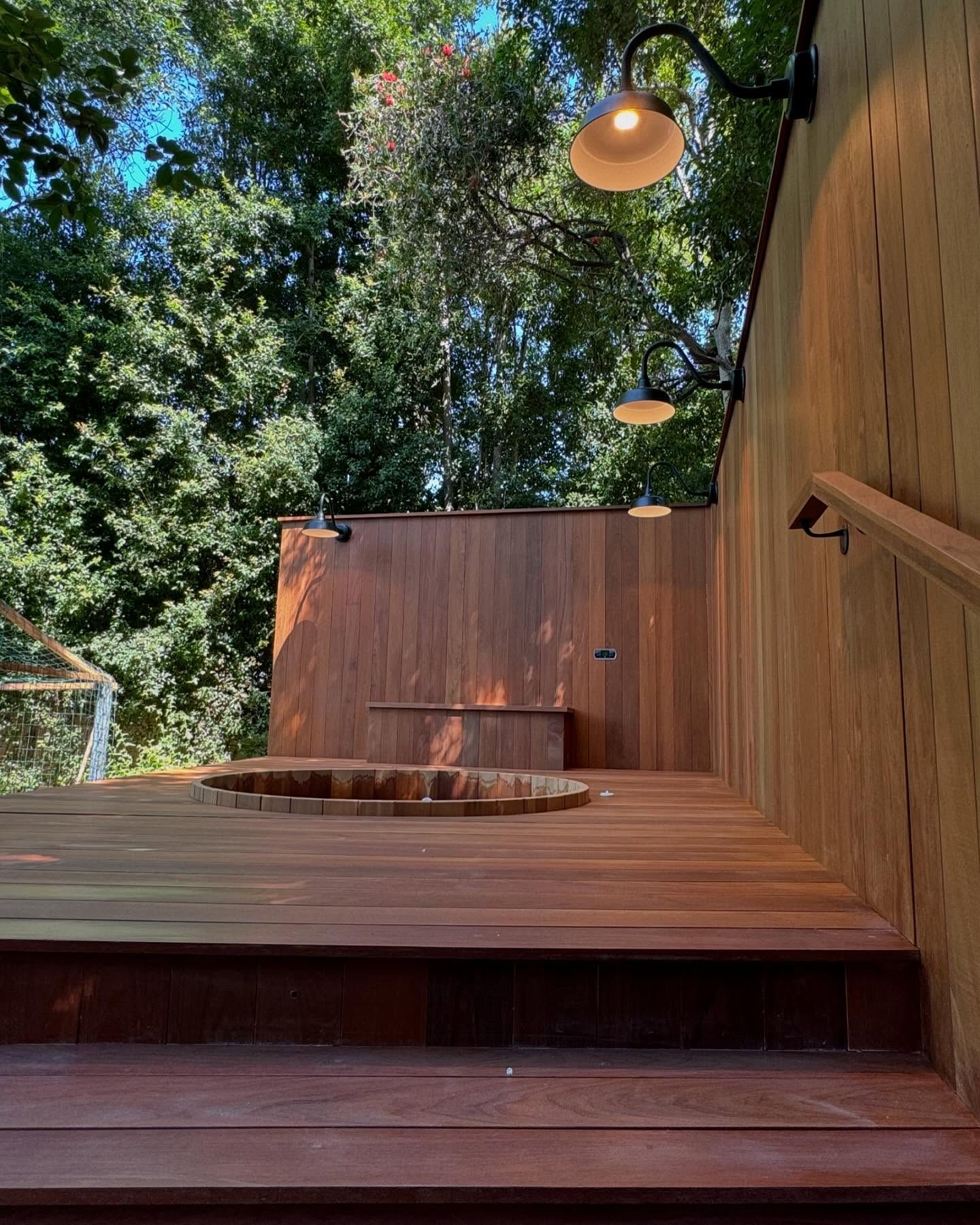 Wooden deck with a built-in hot tub, stairs, and two sconce lights. Forest in the background.