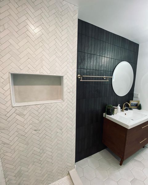 Bathroom with white herringbone tile shower, dark accent wall, and wood vanity with a round mirror.