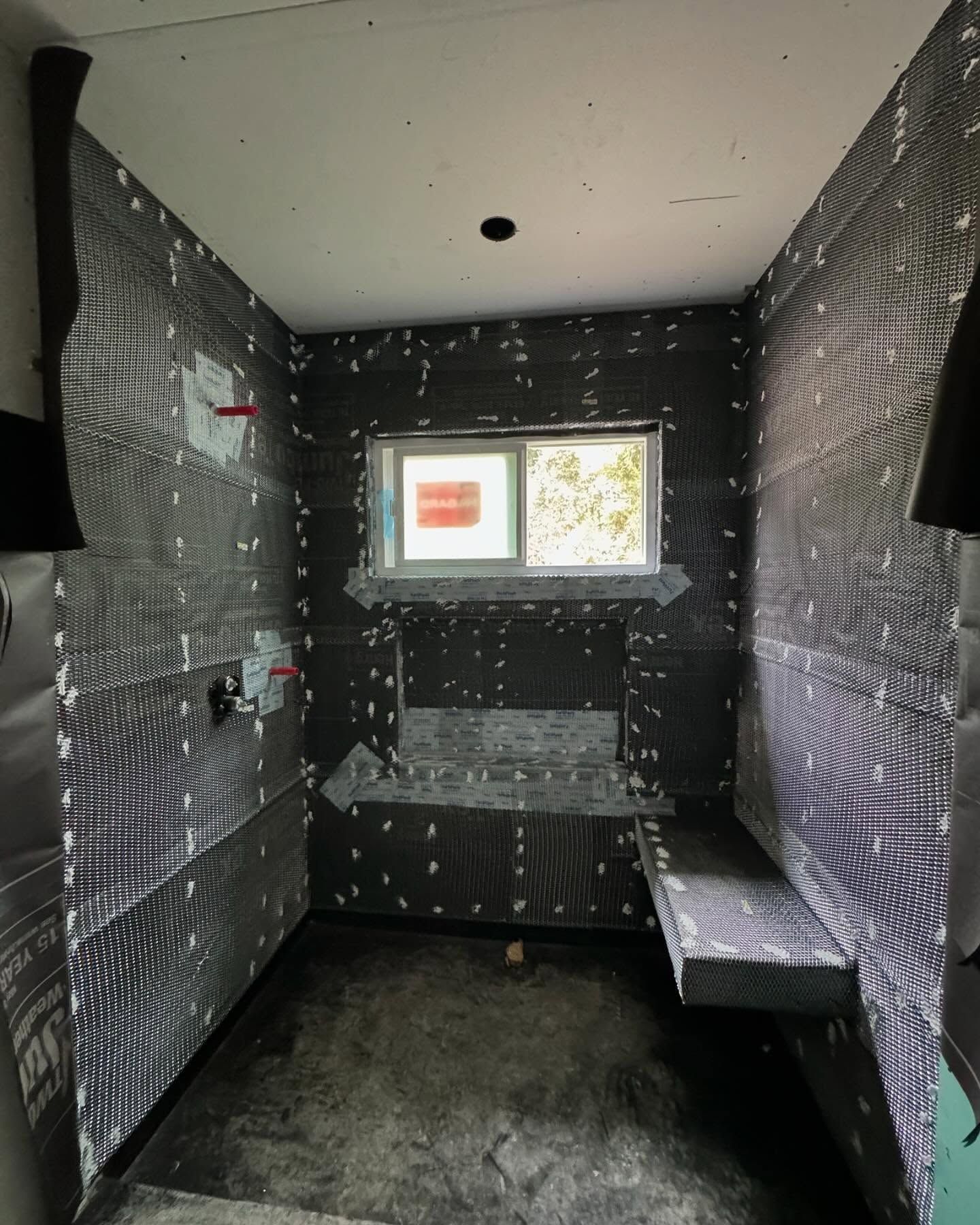 Bathroom under construction, walls covered in black waterproofing membrane, shower bench, small window.
