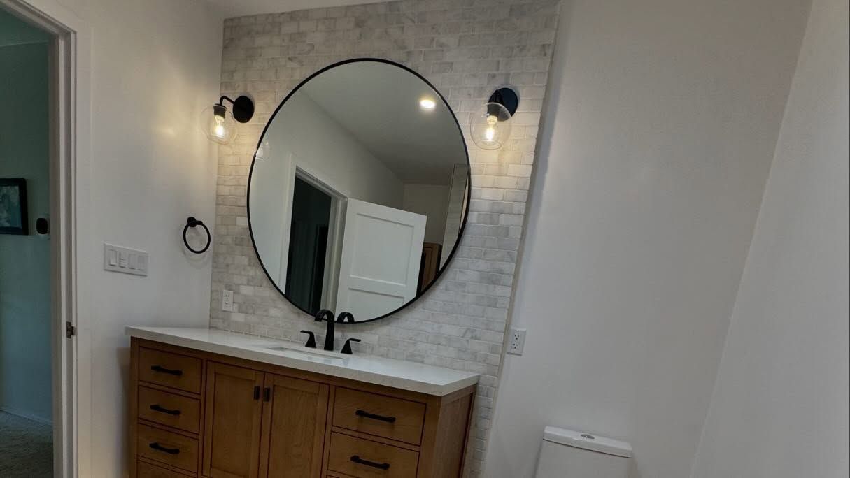 Bathroom with wooden vanity, large round mirror, and tile accent wall.