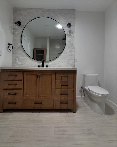 Bathroom with wooden vanity, round mirror, white toilet, and light gray tile.