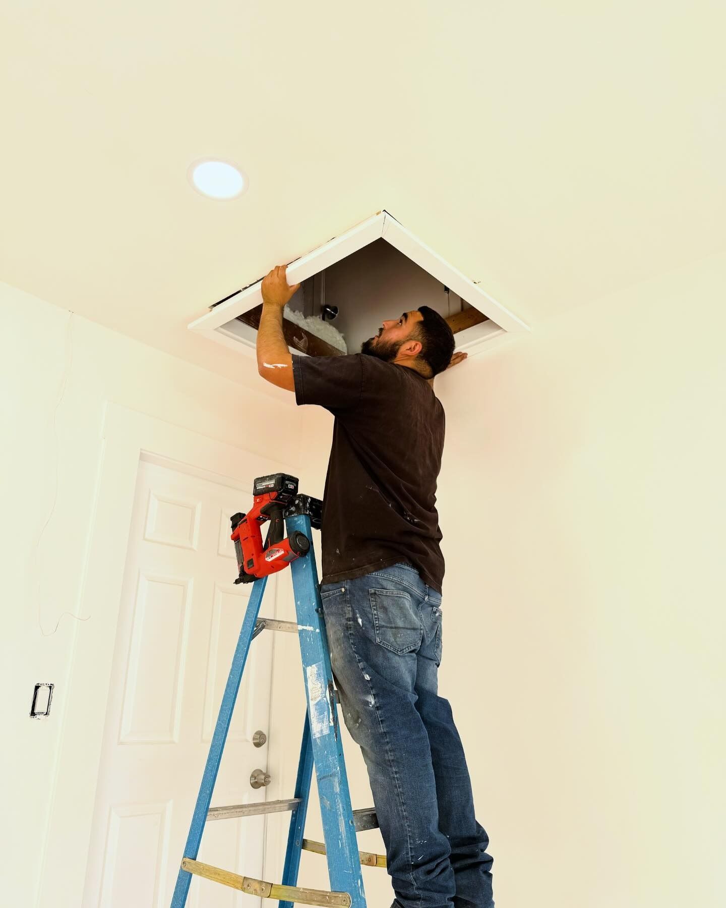 Man on ladder installing an air vent in a white ceiling. He's wearing jeans and a brown shirt in a white room.