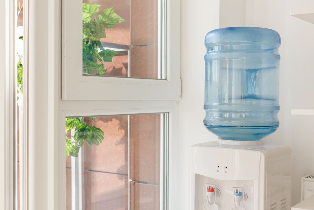 Blue Water Cooler Jug on a Dispenser Beside a Window — A & T Air-Conditioning and Refrigeration in Millbank, QLD