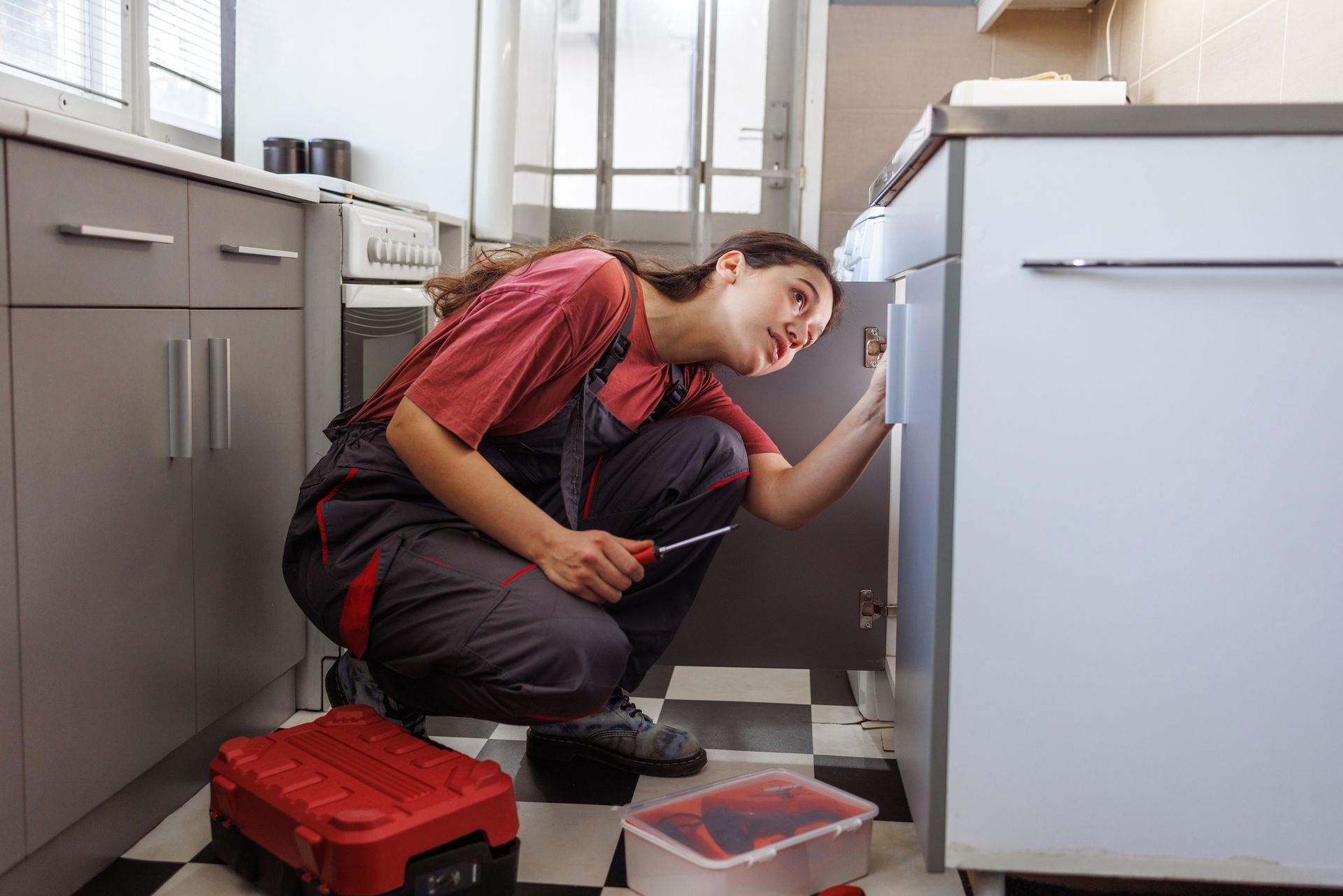 Female technician providing residential plumbing service under kitchen sink with screwdriver.