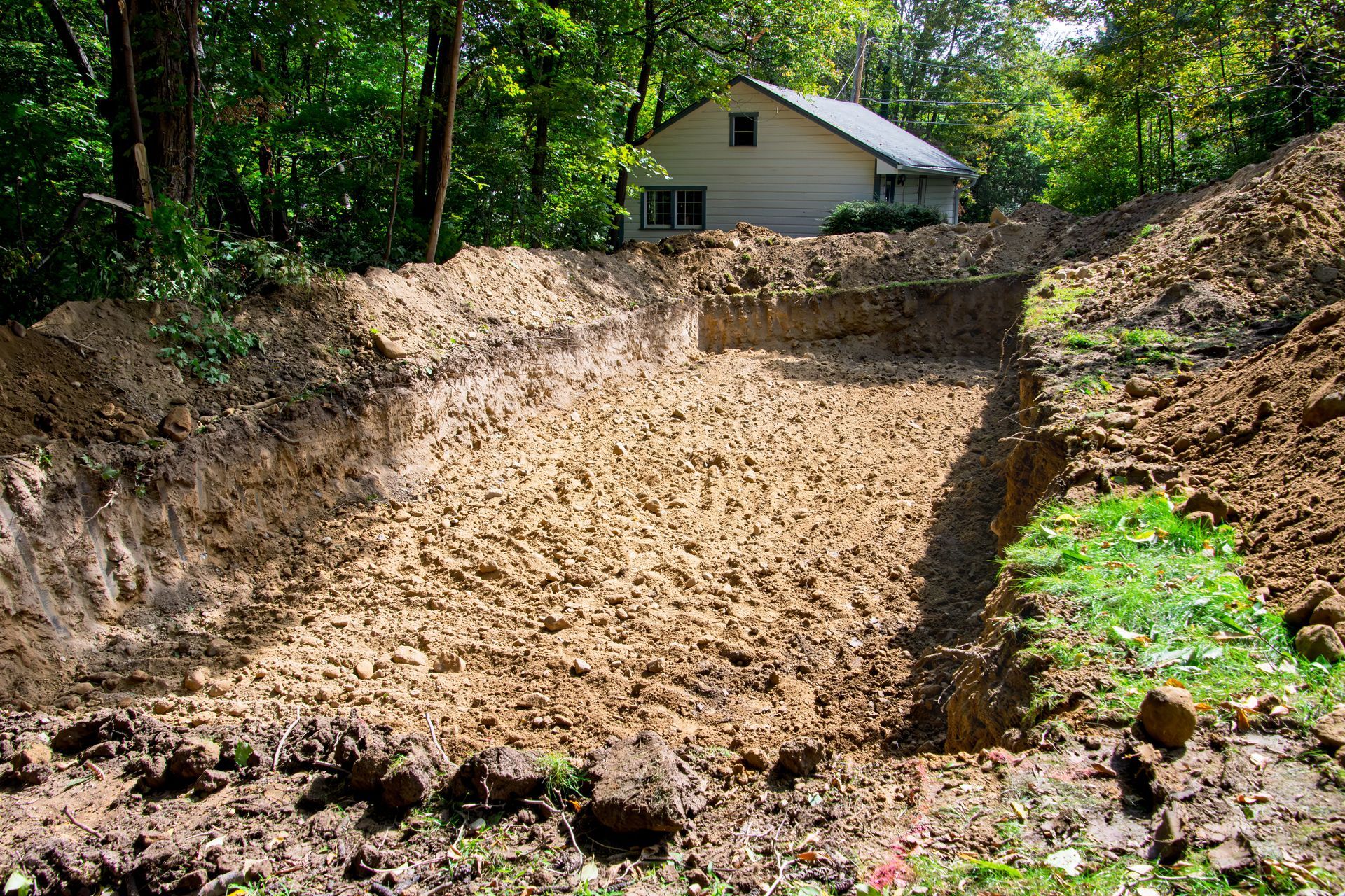 Excavated foundation pit with dirt walls; small house in background, surrounded by trees.