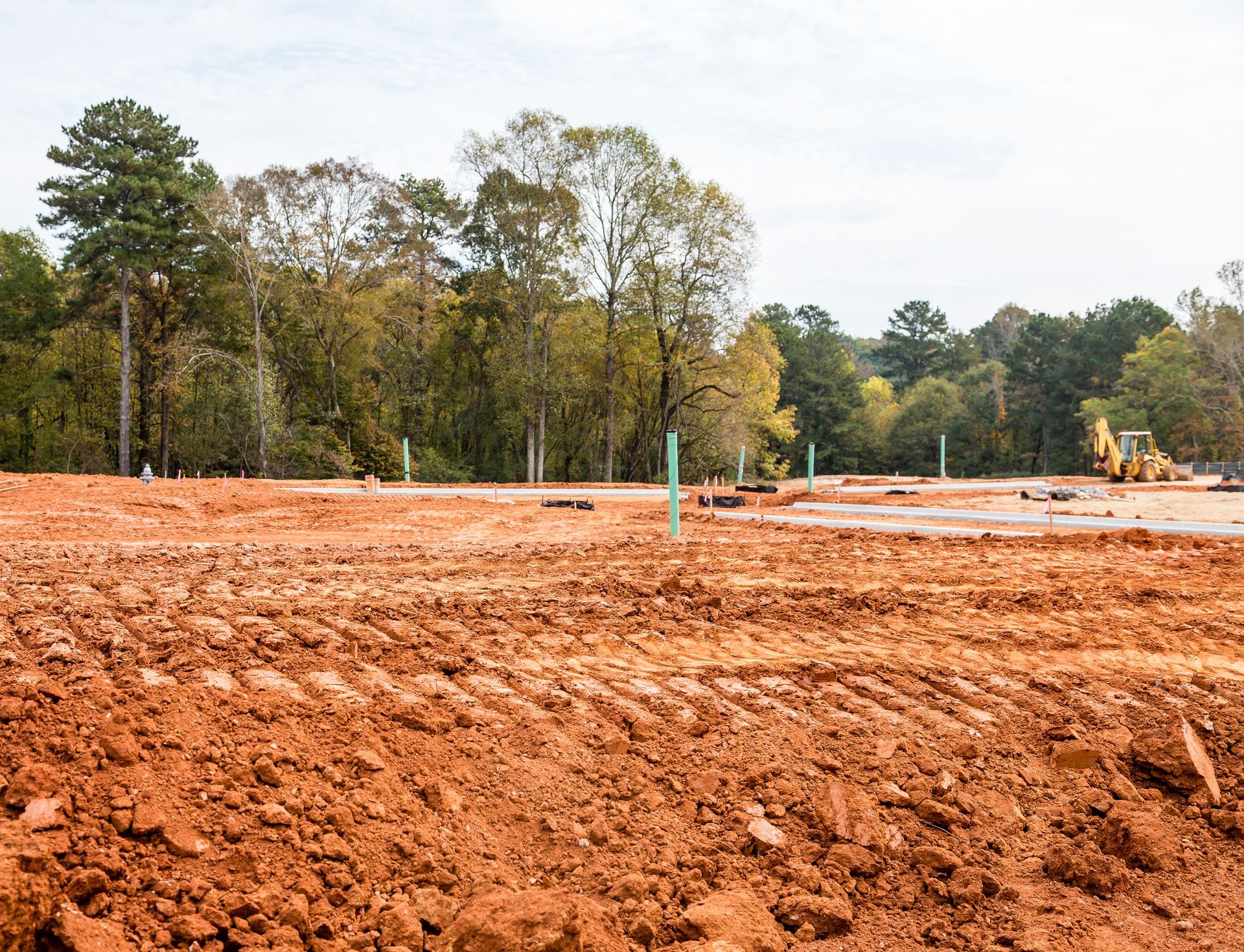 Construction site, red soil in foreground, trees in background, small green pipes and yellow excavator visible.