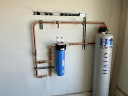 Copper pipes and water filter system mounted on a white wall. Includes a blue filter housing and a silver tank.